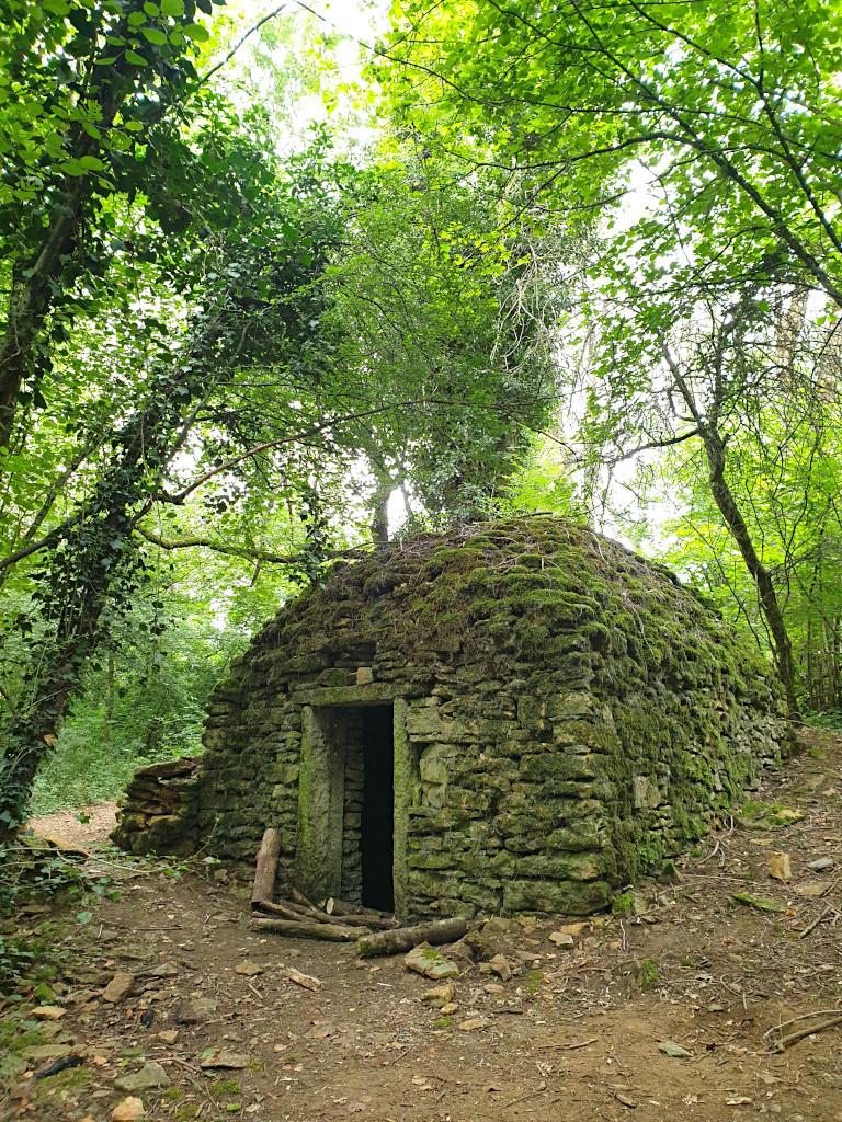 Ancienne cabane en pierres sèches au sommet arrondi couvert de mousse. A l'entrée se trouvent quelques rondins de bois. La cabane est entourée de quelques arbres. Ce type de cabane, appelé caborne, est typique des Monts d'Or lyonnais. 